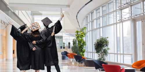 Three recent graduates in caps and gowns celebrating in an office building